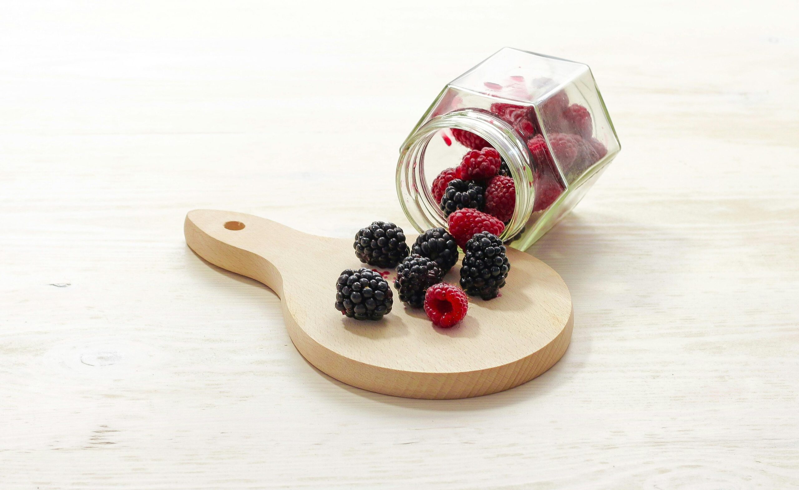 Frutas vermelhas frescas, incluindo amoras e framboesas, derramando de um pote de vidro sobre uma tábua de madeira, ideais para controlar a fome de forma saudável.
