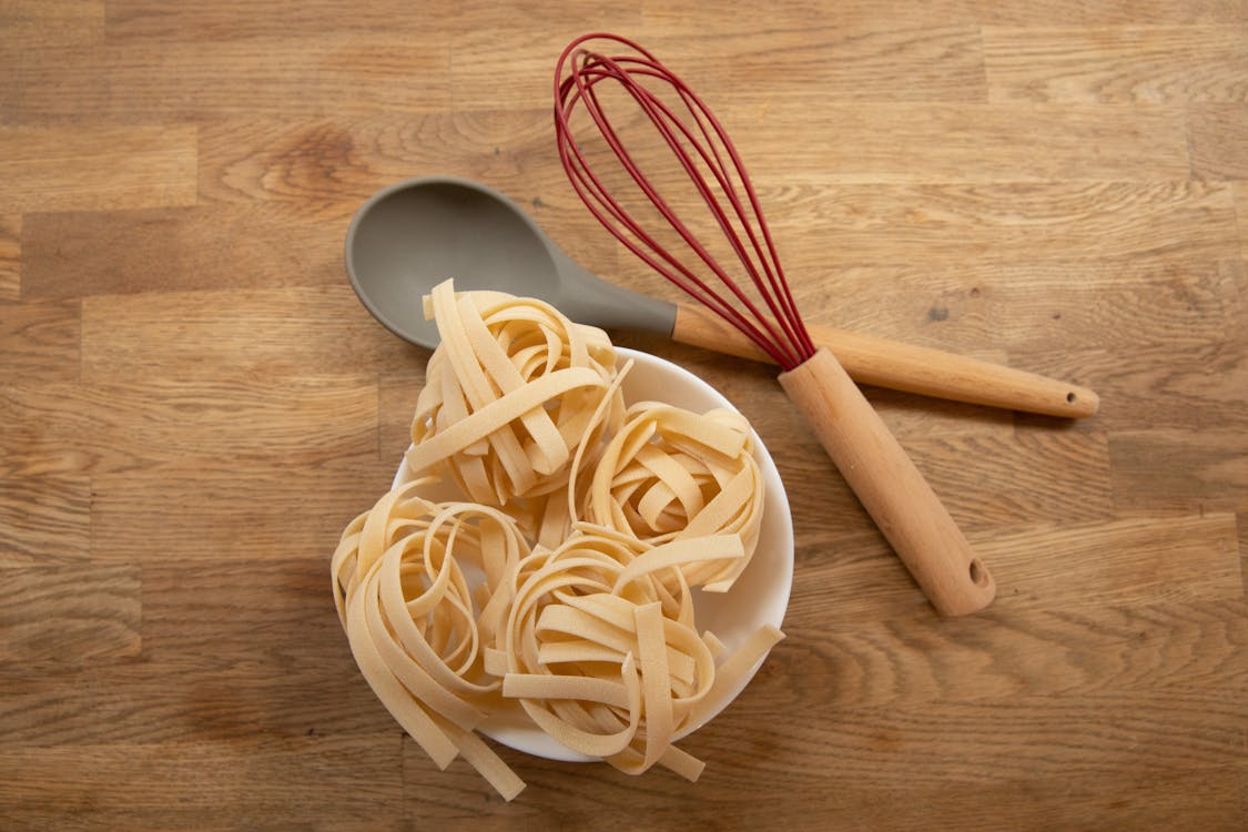 Massa fresca tagliatelle em formato de ninhos dentro de uma tigela branca, acompanhada de colher de silicone e batedor de arame em fundo de madeira. Ingrediente para pratos com carboidratos.