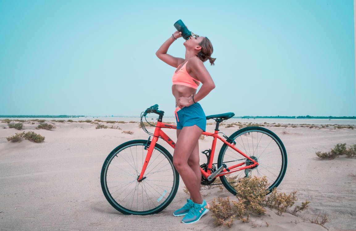 Mulher bebendo água ao lado de uma bicicleta em uma praia ensolarada, vestindo roupas esportivas e tênis, representando um estilo de vida saudável e ativo.