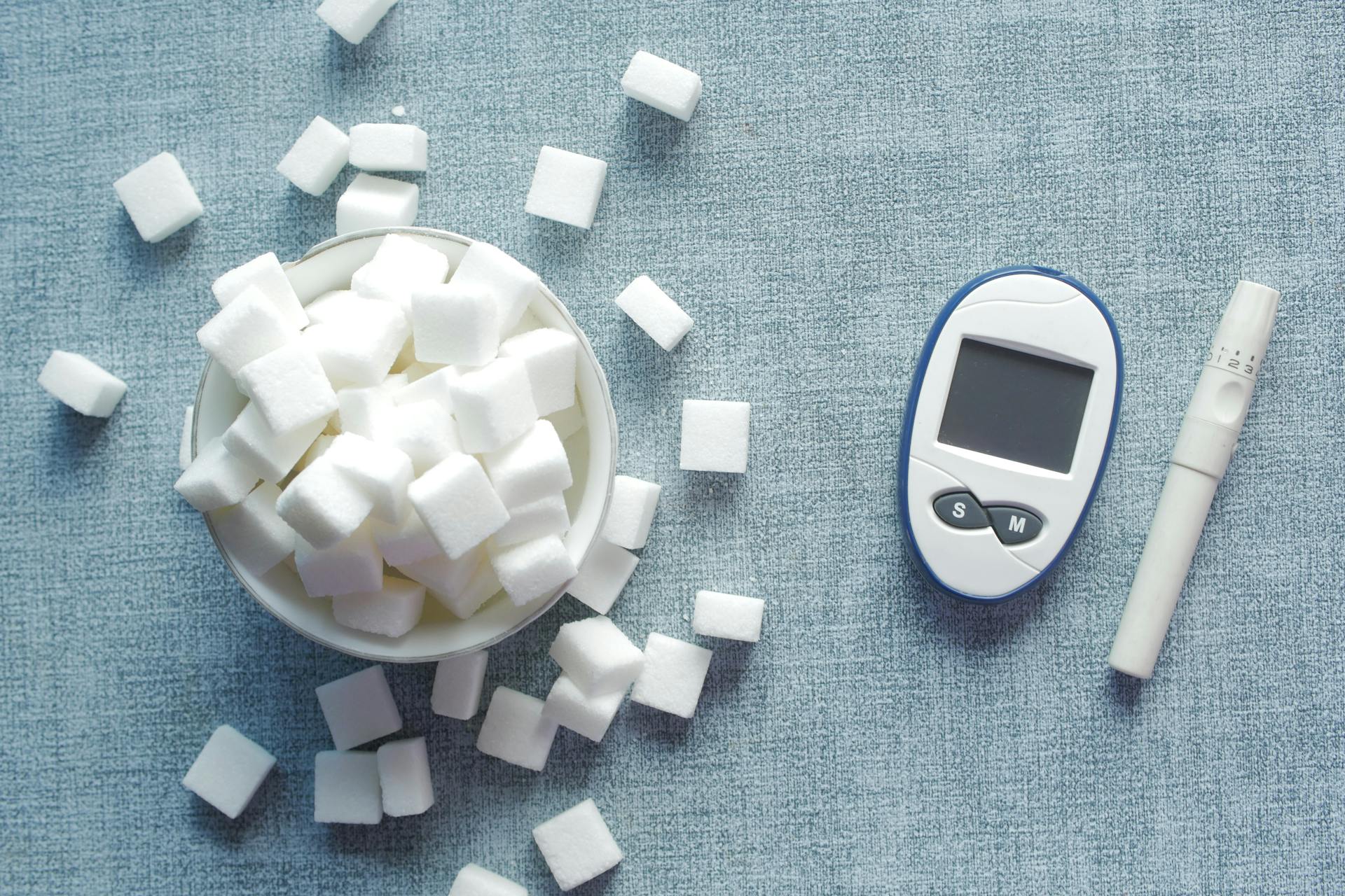 Glicosímetro digital branco e azul ao lado de tigela com cubos de açúcar branco espalhados sobre superfície azul, representando controle de diabetes e monitoramento de glicemia