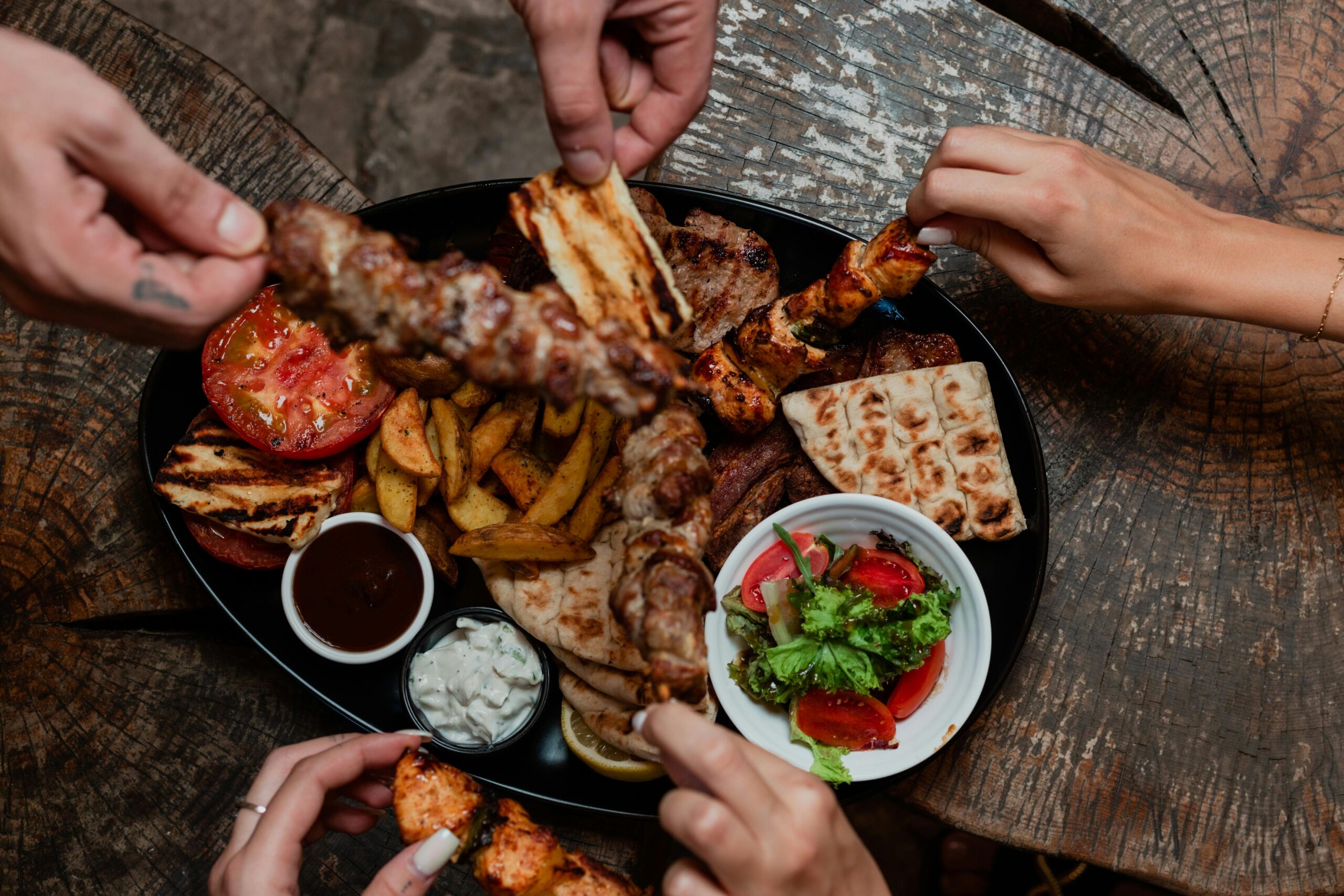 Prato preto com alimentos da dieta mediterrânea incluindo carnes grelhadas, vegetais, pão pita, salada fresca e molhos, sendo compartilhado por várias pessoas em mesa de madeira