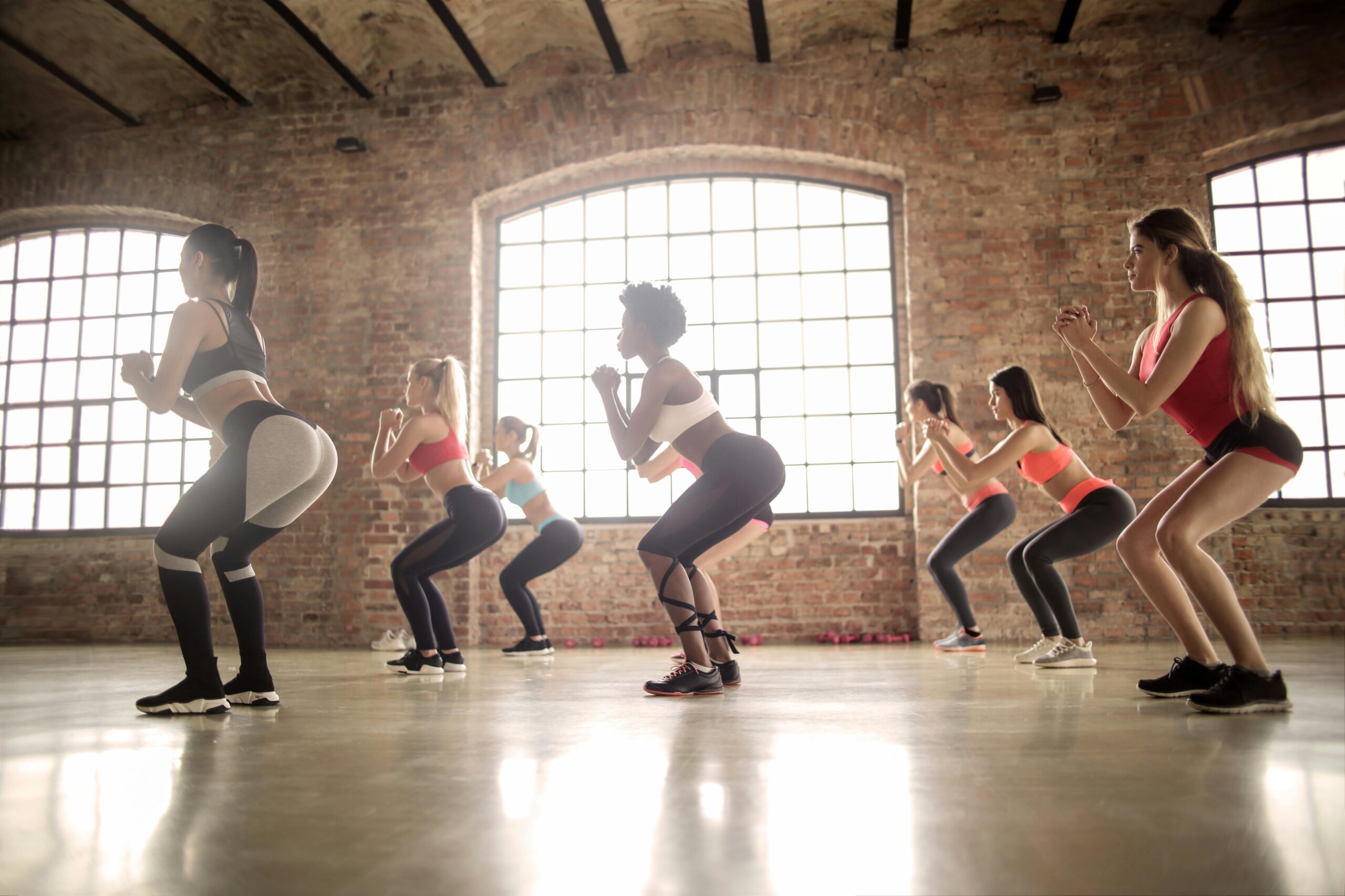 Grupo de mulheres executando agachamentos em academia com parede de tijolo, demonstrando exercícios para glúteos em casa