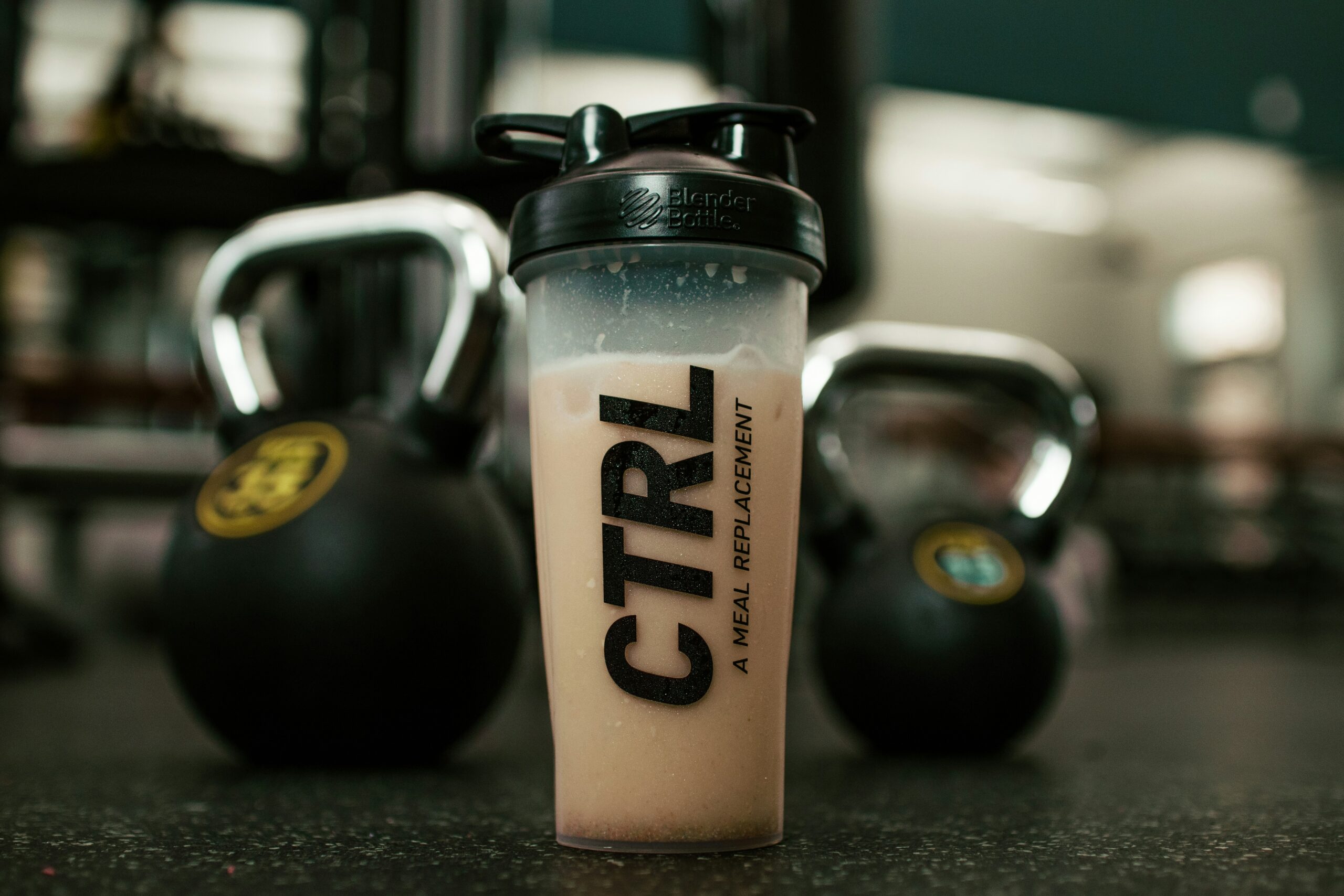 Blender bottle com shake de whey protein ao lado de kettlebells em academia, demonstrando suplementação pós-treino