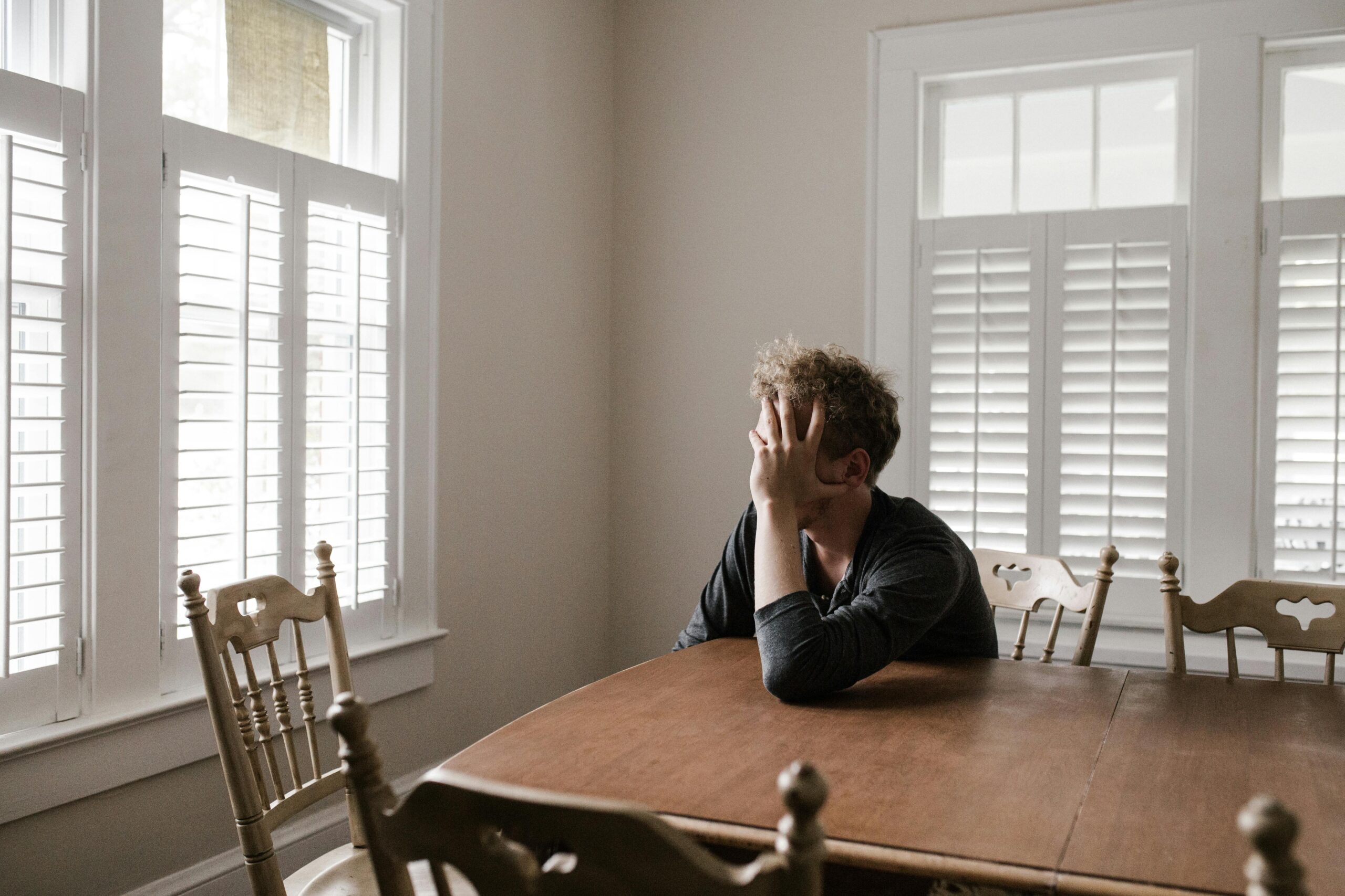Homem jovem com cabeça nas mãos sentado à mesa de madeira em ambiente doméstico iluminado, expressando tristeza e sintomas de depressão que podem ser aliviados com exercícios físicos
