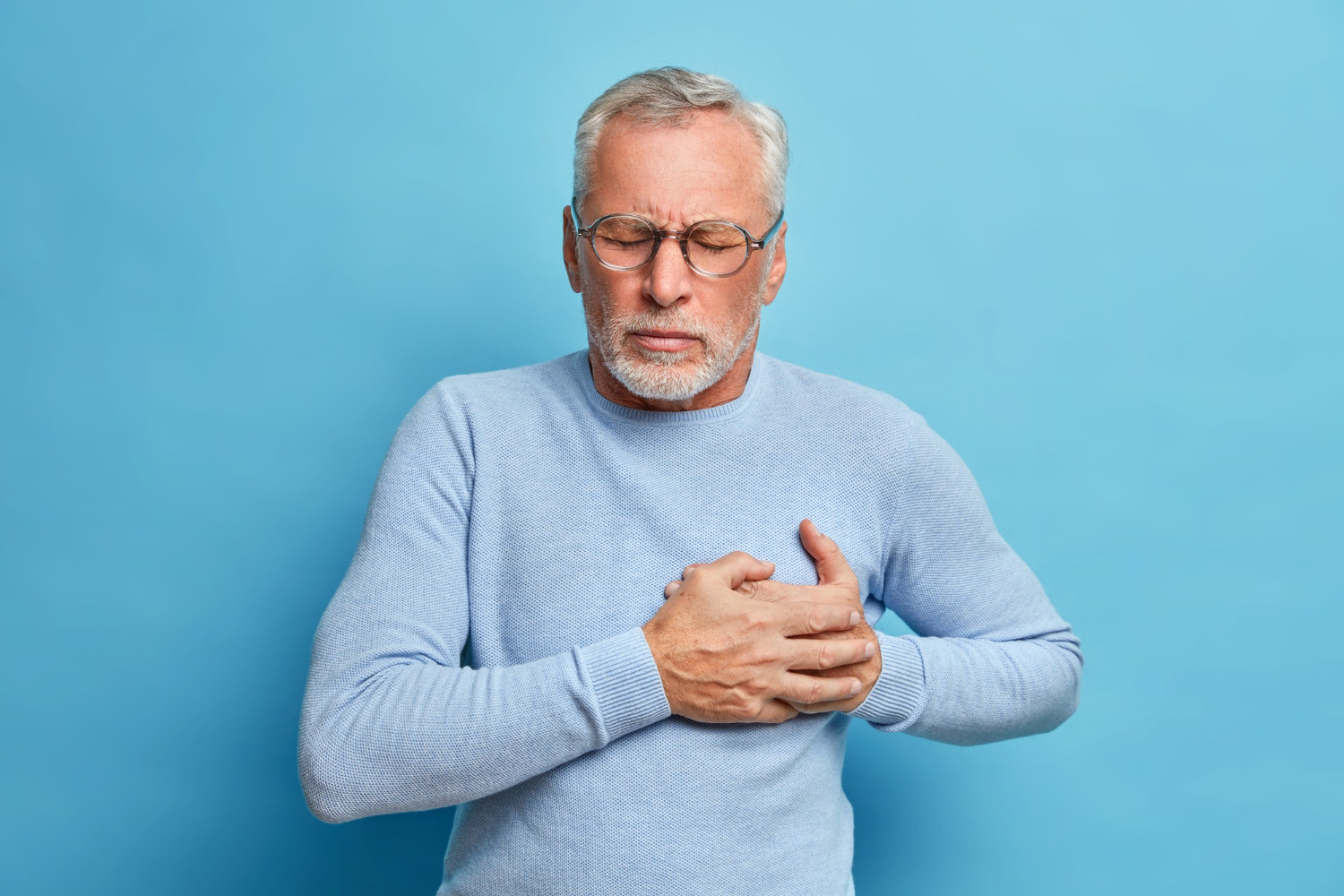 Homem de 60 anos com óculos segurando o peito com as duas mãos, demonstrando desconforto cardíaco, usando suéter azul claro em fundo azul