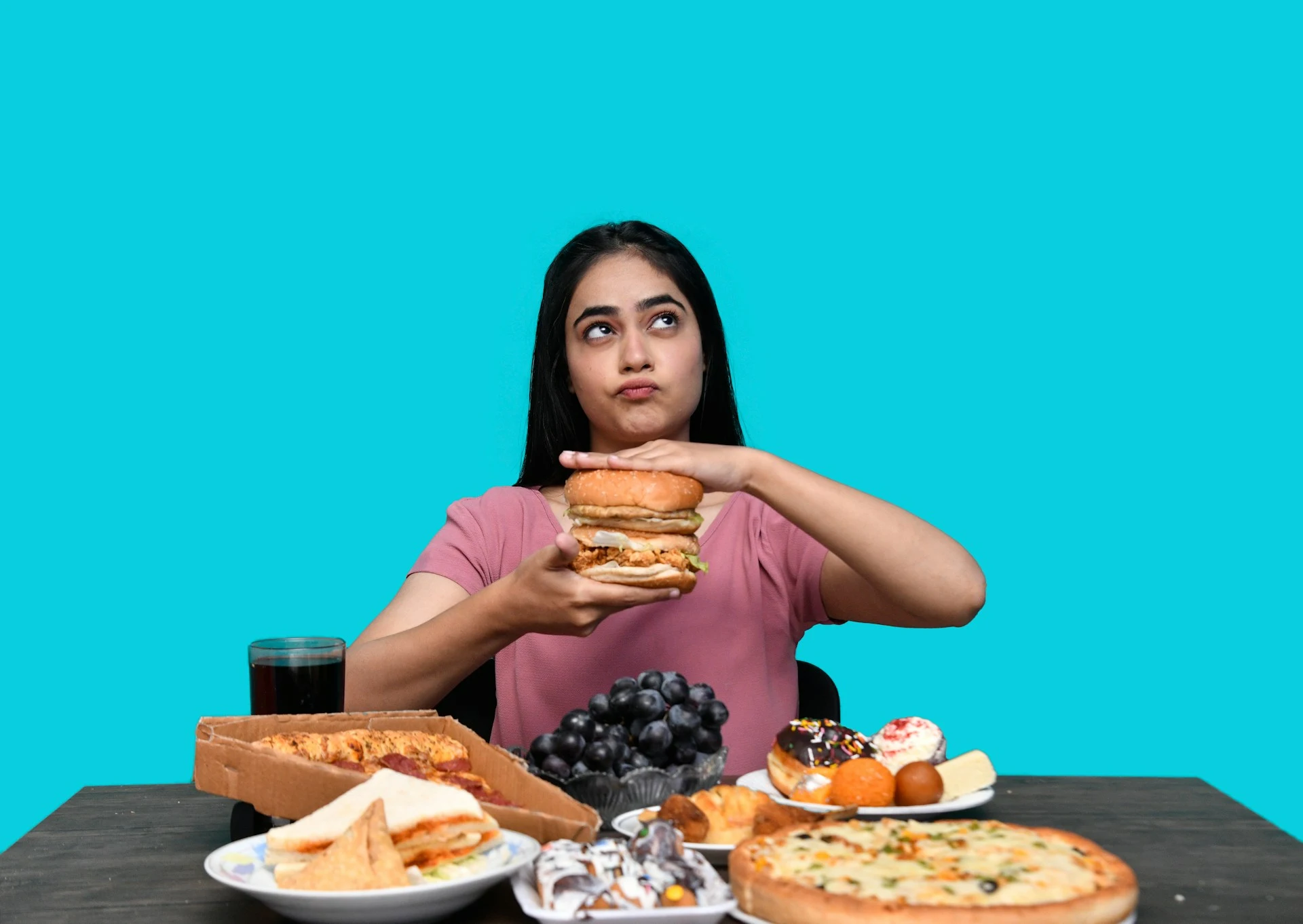 Mulher jovem segurando hambúrguer cercada de alimentos ultraprocessados como pizza, donuts e refrigerante, ilustrando fatores dietéticos que contribuem para síndrome metabólica